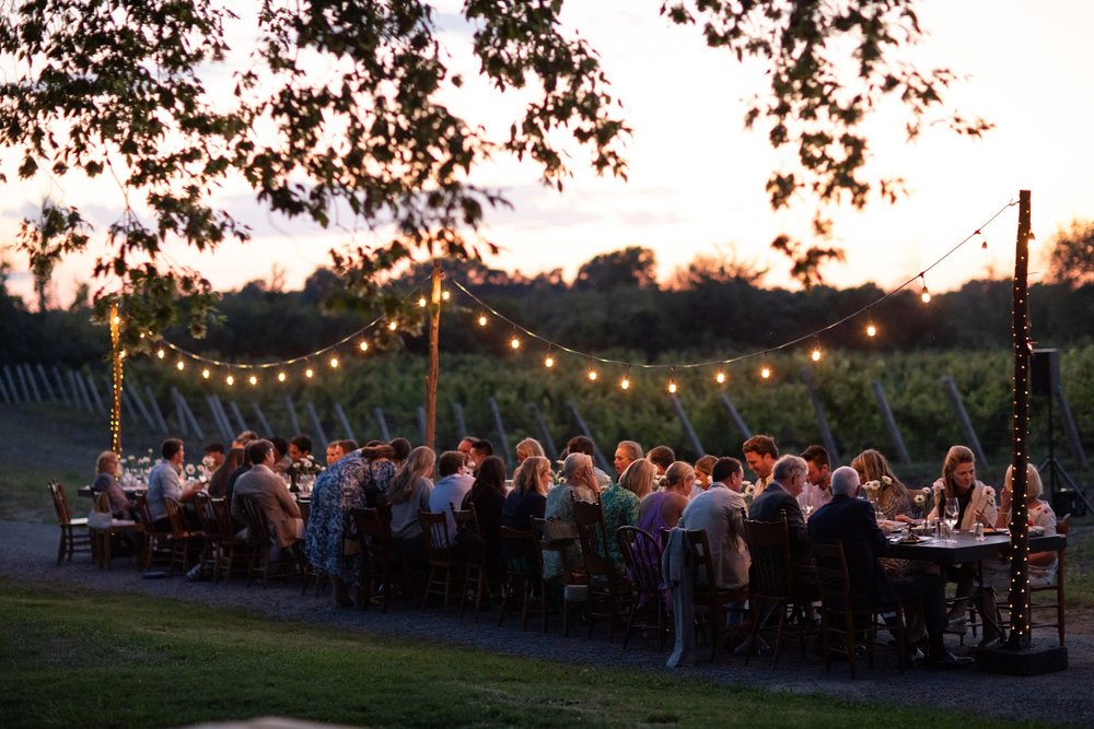 a large group of people dining at an outdoor table at Grange of Prince Edward Estate Winery with fairy lights above them