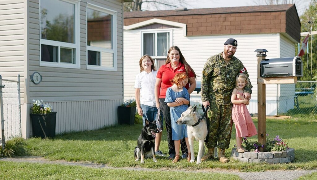 a family with a mother, three children, two dogs and husband in his military uniform stand outside their home