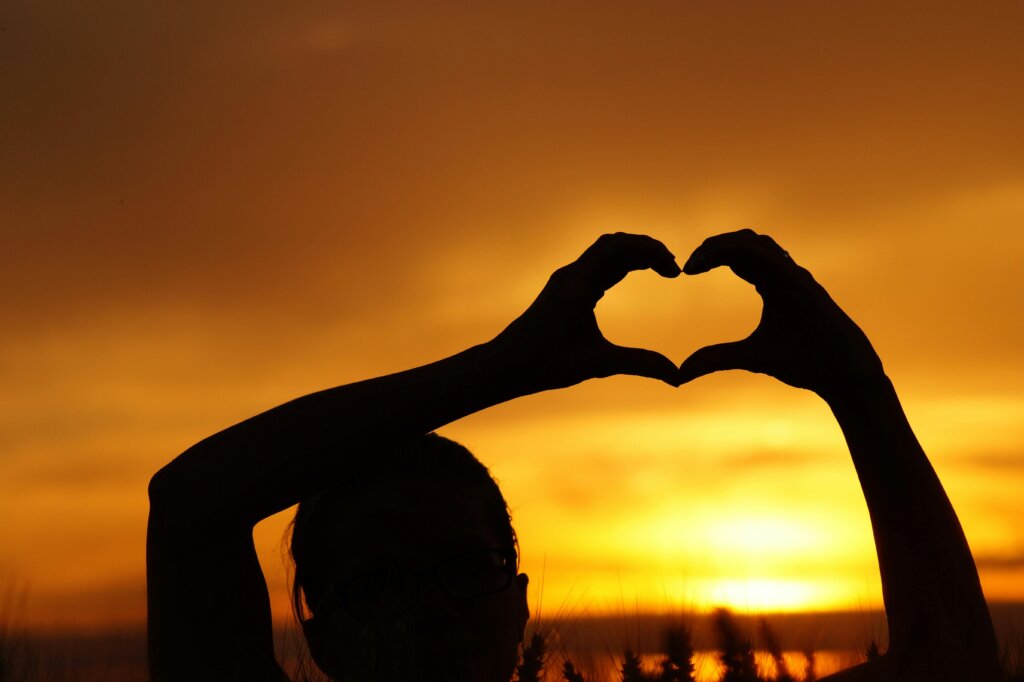  a woman making a heart shape with her hands in front of an orange sunset